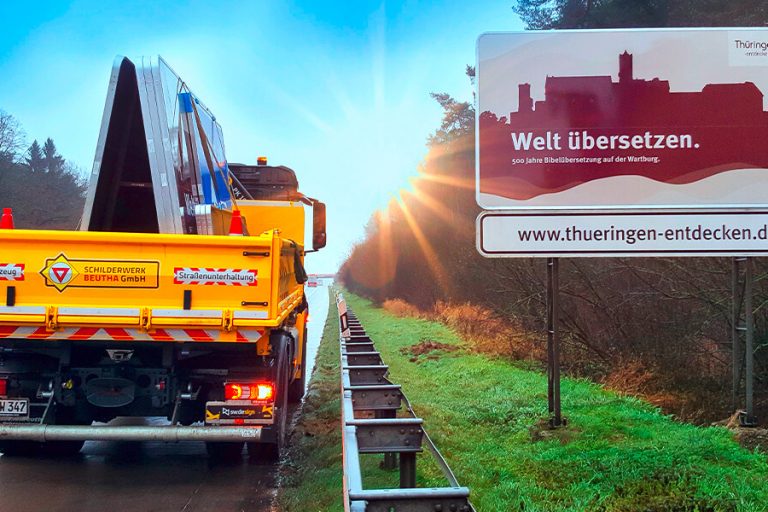 Foto einer touristischen Unterrichtstafel mit der Aufschrift: Welt übersetzen. 500 Jahre Bibelübersetzung auf der Wartburg, www.thueringen-entdecken.de Neben dem Schild befindet sich ein Baustellenfahrzeug mit neuer Folierung der Schilderwerk-Beutha GmbH