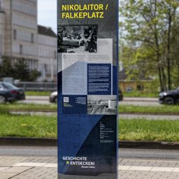 Fotografie unseres Produktes der Stele Regulus am Standort Falkeplatz in Chemnitz. Die Stele befindet sich im Vordergrund, im Hintergrund sind leicht verschwommen fahrende Autos zu sehen.