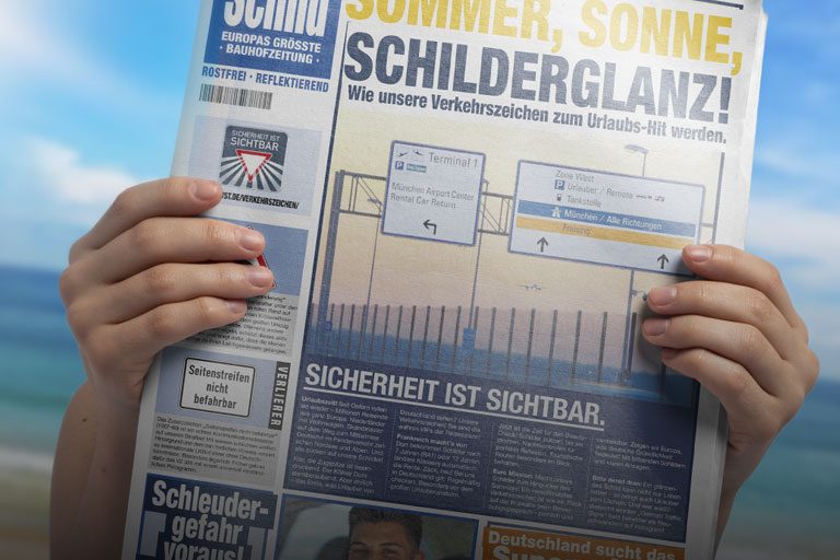 Zwei Hände halten die aktuelle Schild-Zeitung hoch. Im Hintergrund ist verschwommen Strand und Meer zu sehen - passend zum Titelthema der SCHILD "Sommer, Sonne, Schilderglanz".