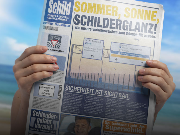 Zwei Hände halten die aktuelle Schild-Zeitung hoch. Im Hintergrund ist verschwommen Strand und Meer zu sehen - passend zum Titelthema der SCHILD "Sommer, Sonne, Schilderglanz".
