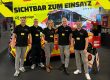 sw/de/sign Stand auf der Florian 2025 mit foliertem Auto und Rückwand mit Feuerwehrwagen und Aufschrift: Sichtbar zum Einsatz. Davor stehen fünf Mitarbeiter des sw/de/sign Teams.