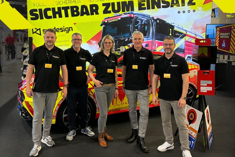 sw/de/sign Stand auf der Florian 2025 mit foliertem Auto und Rückwand mit Feuerwehrwagen und Aufschrift: Sichtbar zum Einsatz. Davor stehen fünf Mitarbeiter des sw/de/sign Teams.