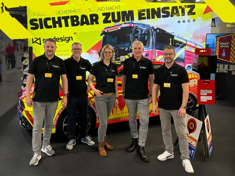 sw/de/sign Stand auf der Florian 2025 mit foliertem Auto und Rückwand mit Feuerwehrwagen und Aufschrift: Sichtbar zum Einsatz. Davor stehen fünf Mitarbeiter des sw/de/sign Teams.