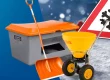 Grauer Streugutbehälter mit orangem Deckel, Schneeschaufel und gelbem Streuwagen vor Schneeflocken-Verkehrsschild