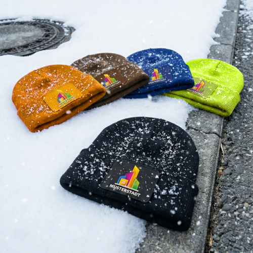 Textiveredelung_Mützenaktion_Logo_verschiedene Farben Mehrere Strickmützen in verschiedenen Farben liegen im Schnee auf einem Bordstein