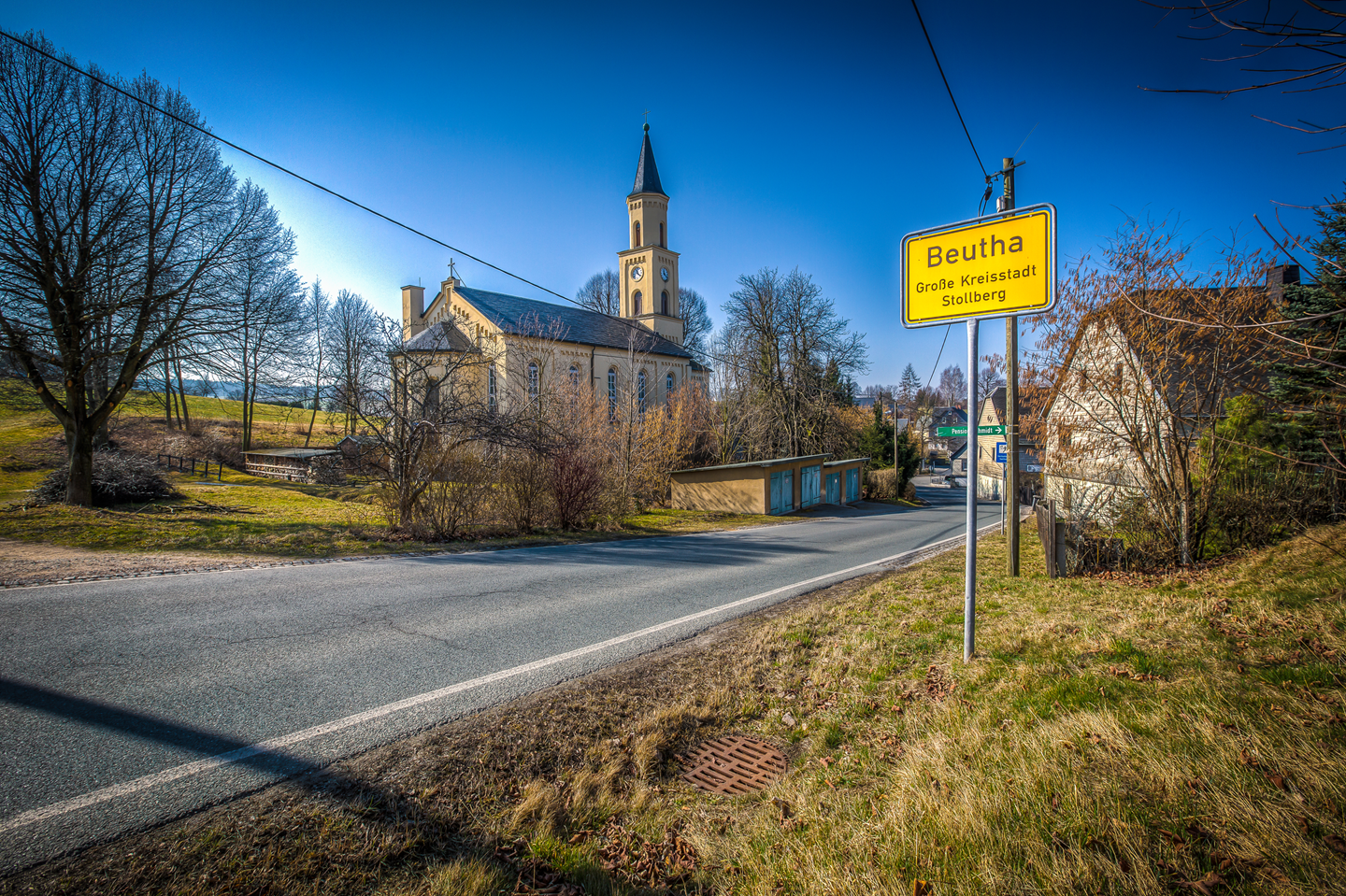 Gemeinde_Beutha_mit Ortstfafel Gelbes Ortsschild „Beutha, Große Kreisstadt Stollberg“ an einer Straße, daneben eine Kirche und Wohnhäuser in einer ländlichen Umgebung bei klarem Himmel.