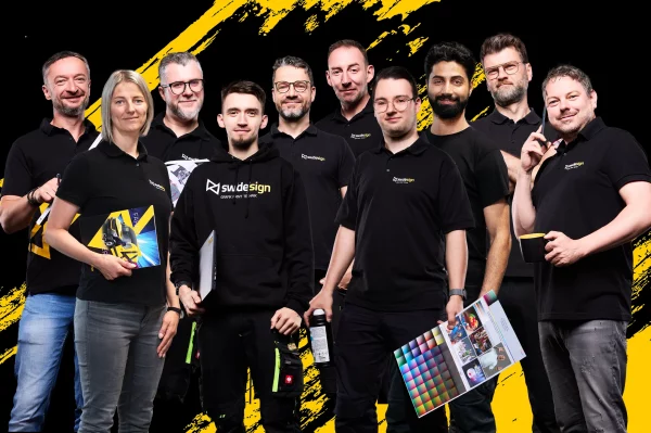 Teamfoto von elf Mitarbeitenden eines Grafik- und Werbetechnikunternehmens mit Arbeitsmaterialien vor schwarzem Hintergrund mit gelben Grafikelementen