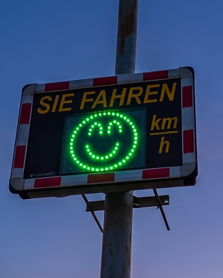 Geschwindigkeitsanzeige mit Smiley zur Verkehrsberuhigung bei Nacht Geschwindigkeitsanzeige mit grünem Smiley bei Nacht an einem Mast in einer Ortschaft. Das Display motiviert Verkehrsteilnehmer zur Einhaltung der zulässigen Geschwindigkeit.
