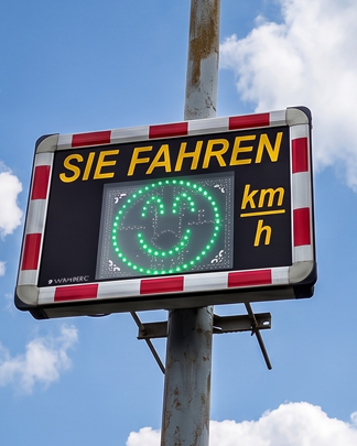 Geschwindigkeitsanzeige mit Smiley zur Verkehrsberuhigung bei Tag Geschwindigkeitsanzeige mit grünem Smiley an einem Mast in einer Ortschaft. Das Display motiviert Verkehrsteilnehmer zur Einhaltung der zulässigen Geschwindigkeit.