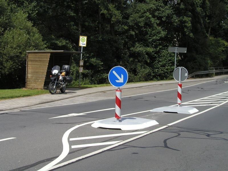 Fahrbahnteiler mit Verkehrszeichen zur sicheren Verkehrsführung Fahrbahnteiler mit Leitbaken und blauem Verkehrszeichen zur Verkehrsführung auf einer Straße.