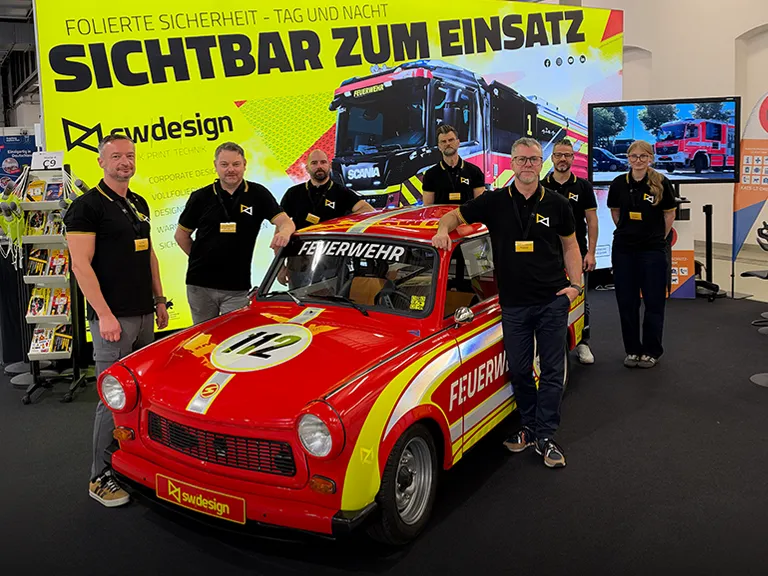 Sechs Teammitglieder von SW Design am Messestand der FLORIAN 2026 mit einem rot-gelben Feuerwehr-Trabant.